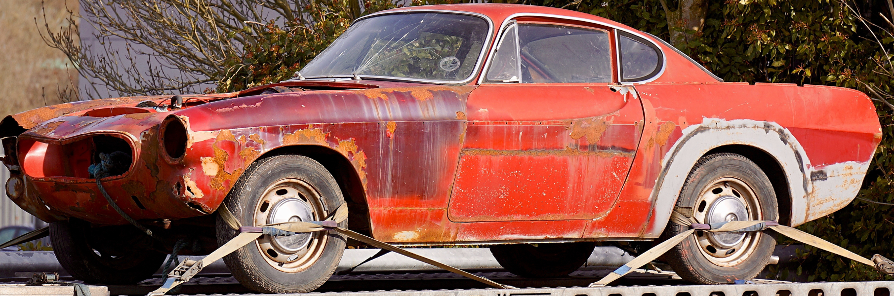 Old orange junk car that is rusting. The car is strapped to the platform on the back of a truck.